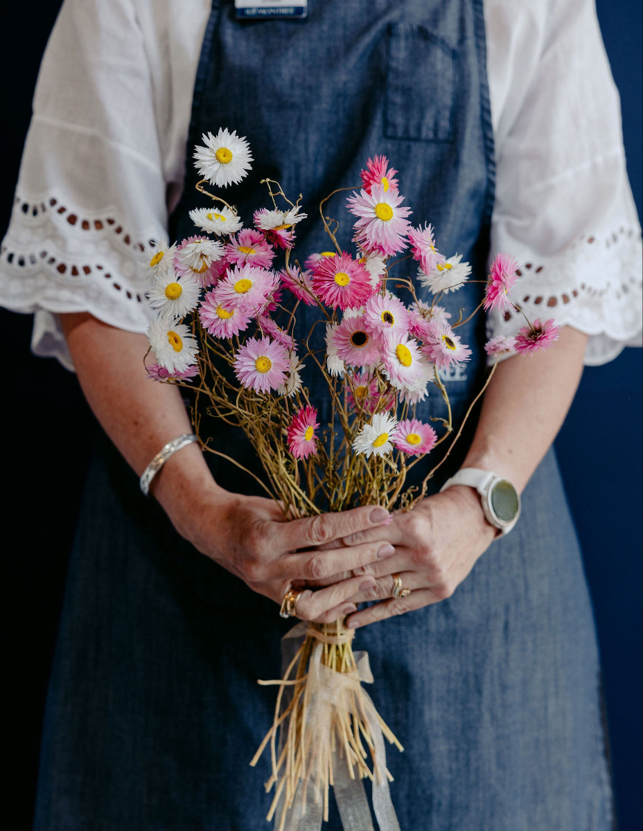 A person wearing a white eyelet blouse and a blue denim apron holds a small bouquet of pink, white, and yellow daisy-like flowers with dried stems and ribbon ties against a dark background.