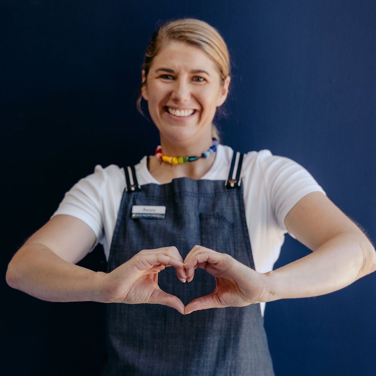 A smiling woman with blonde hair tied back posing with her hands forming a heart shape in front of her.