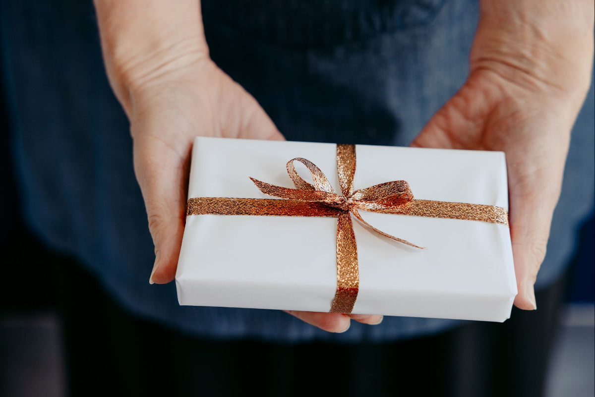 A close-up of a person's hands holding a white gift box wrapped with a glittery copper-brown ribbon and bow, against a dark blue denim shirt background.