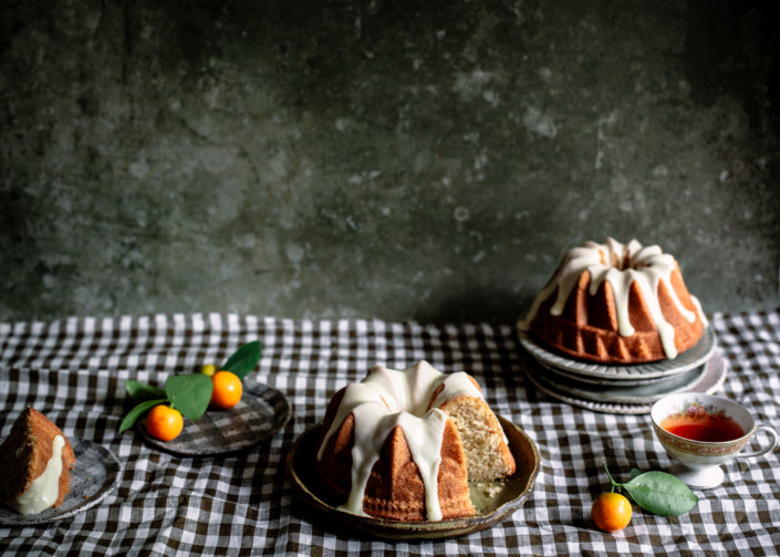 Bundt cakes with white icing on a checkered tablecloth with oranges and a teacup.