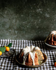 Bundt cakes with white icing on a checkered tablecloth with oranges and a teacup.